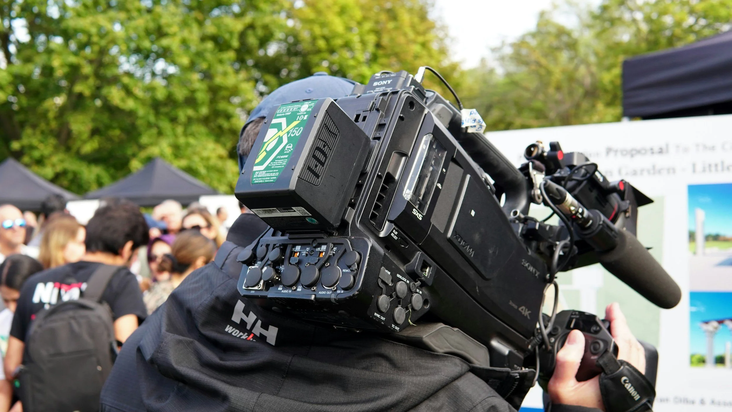 A person operating a large professional video camera in an outdoor event with a crowd and trees in the background.