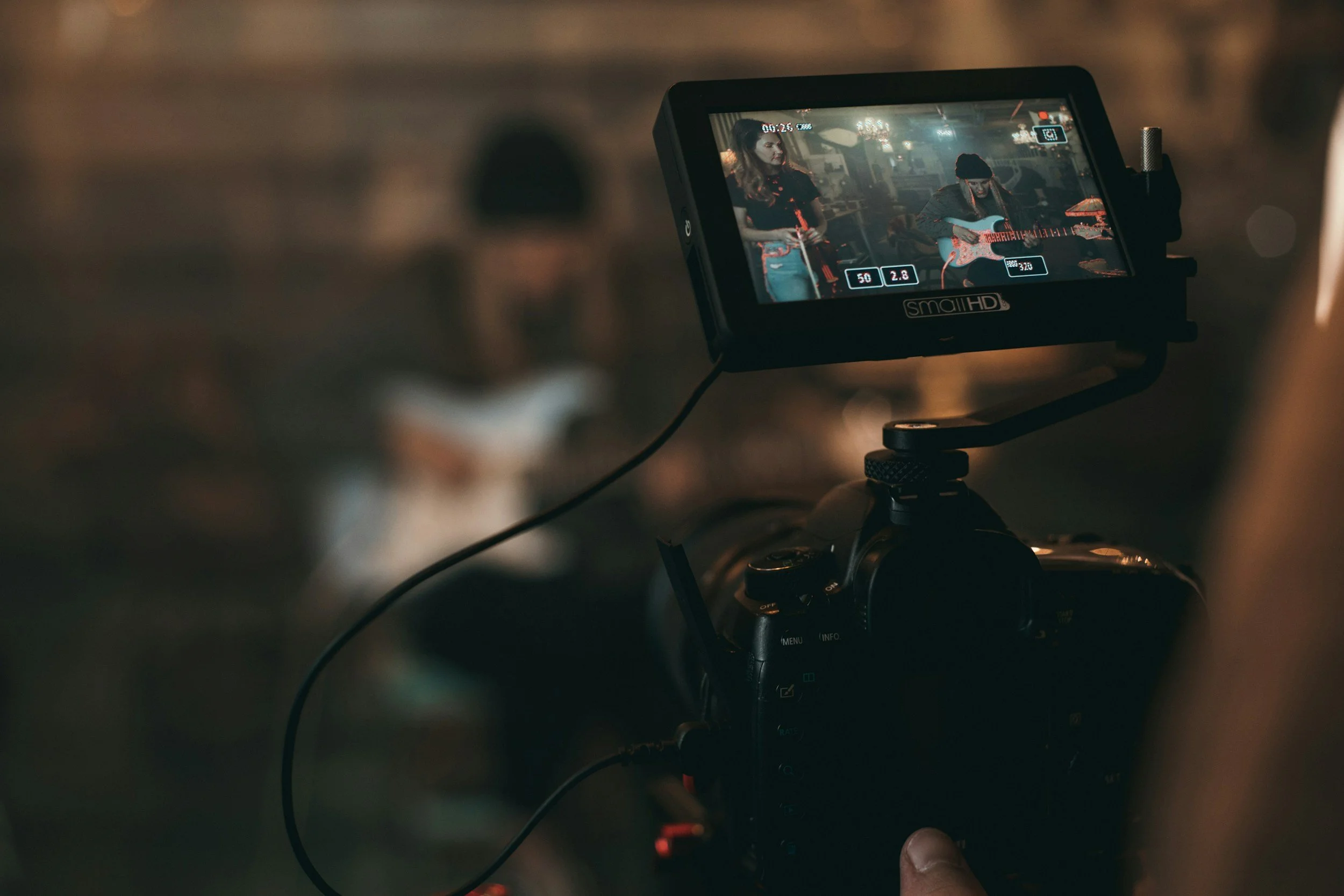 A camera monitor showing two women, one with long curly hair wearing a black top, and the other with straight hair wearing a beanie, playing guitars in a dimly lit indoor scene.