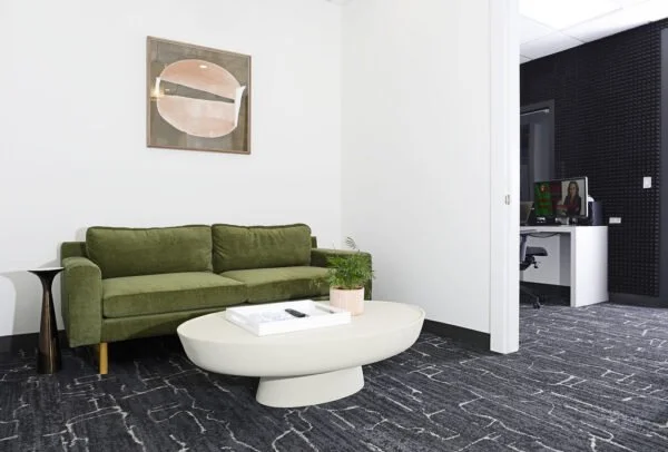 Living room with a green sofa, a white oval coffee table with a plant and book on it, a small side table, and wall art, leading to a workspace with a white desk and monitor.