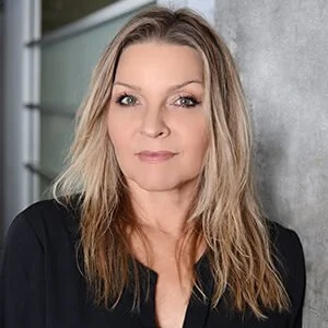 A woman with shoulder-length blonde hair and neutral makeup, wearing a black top, standing indoors with a metal garage door and gray wall in the background.