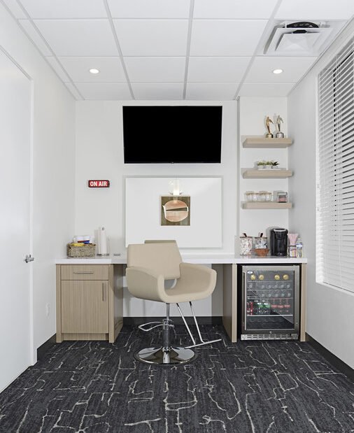 A small radio station or office with a beige chair in front of a desk, a TV mounted on the wall, and a mini fridge below the desk with drinks inside. There are shelves on the right wall holding small items, and an "On Air" sign on the left wall.