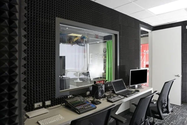 A recording or radio studio with a soundproof window, audio equipment, computers, and chairs.