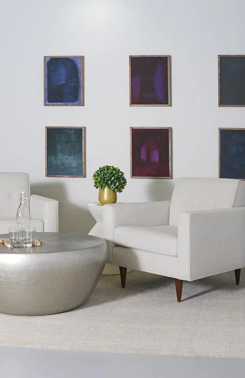 Modern living room with white armchairs, a round metallic coffee table with water bottles, a small white side table with a green plant in a gold pot, and six abstract paintings on the white wall.