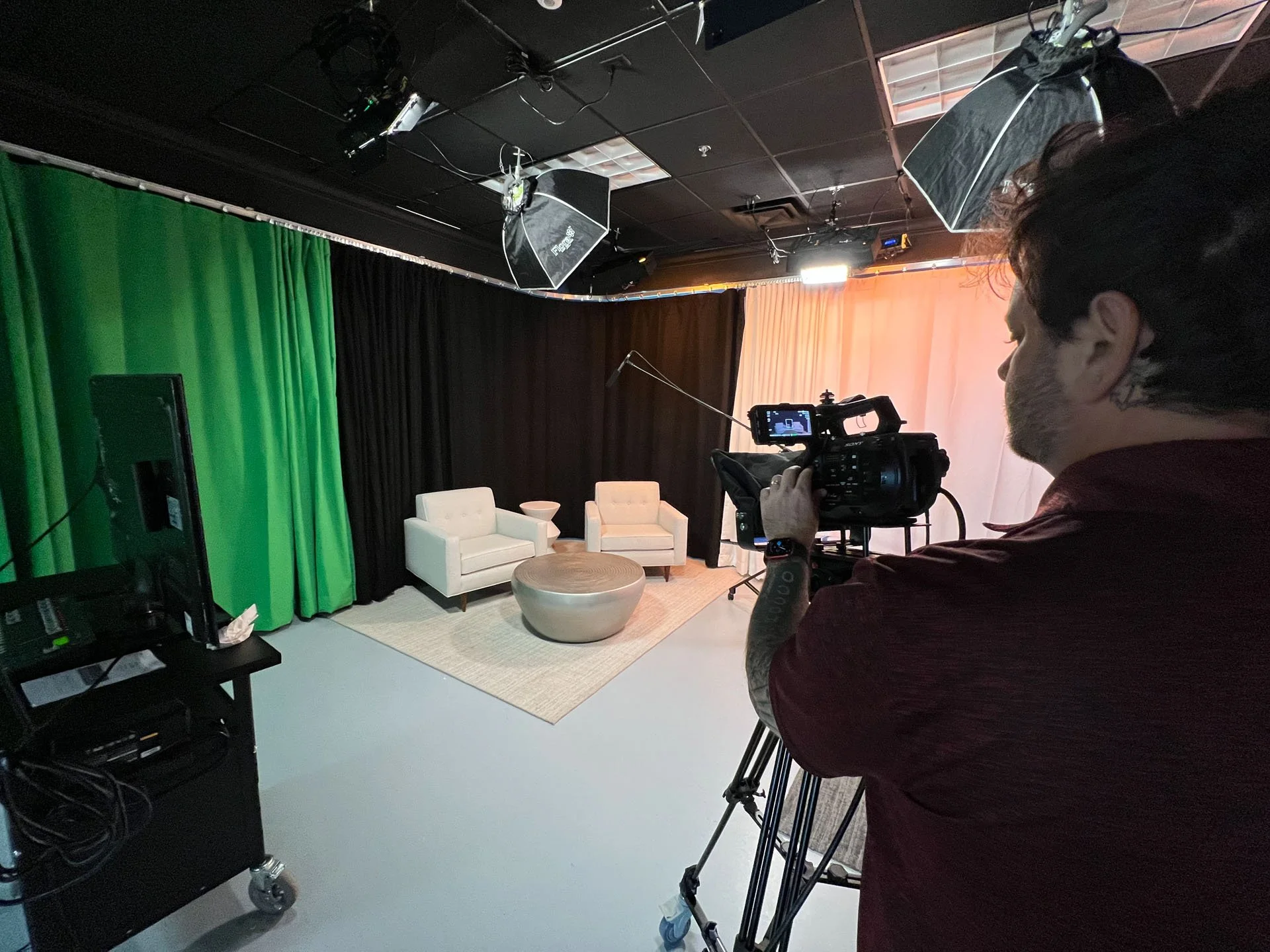 A person filming a television set with a film camera in a studio. The studio has a setup with a sofa, chairs, and curtains, along with lighting equipment and green screens.