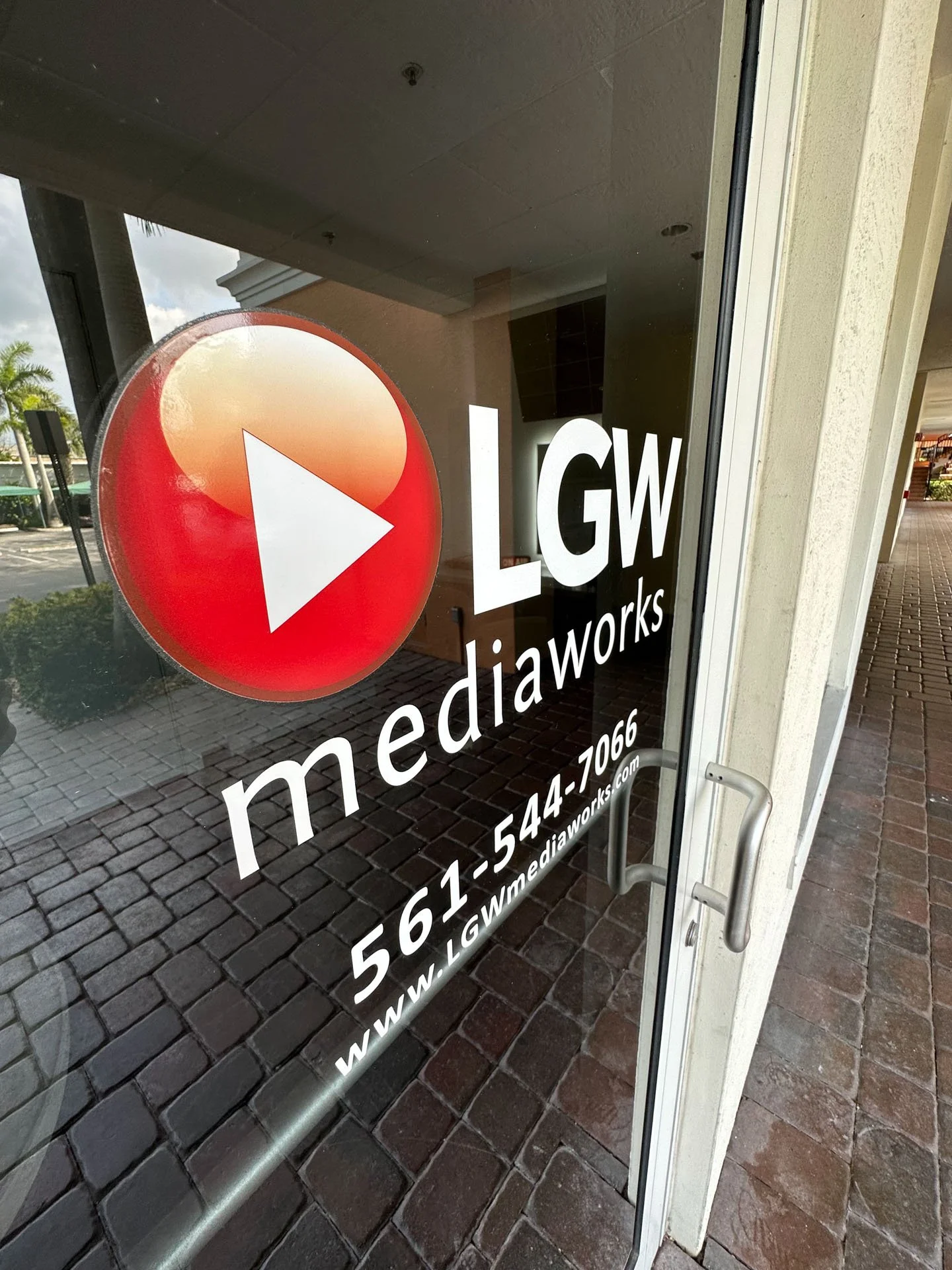 Storefront window with a logo resembling a red circle with a white play button, displaying the text 'LGW media' and contact information. Reflection of outside area with palm trees and sidewalk.