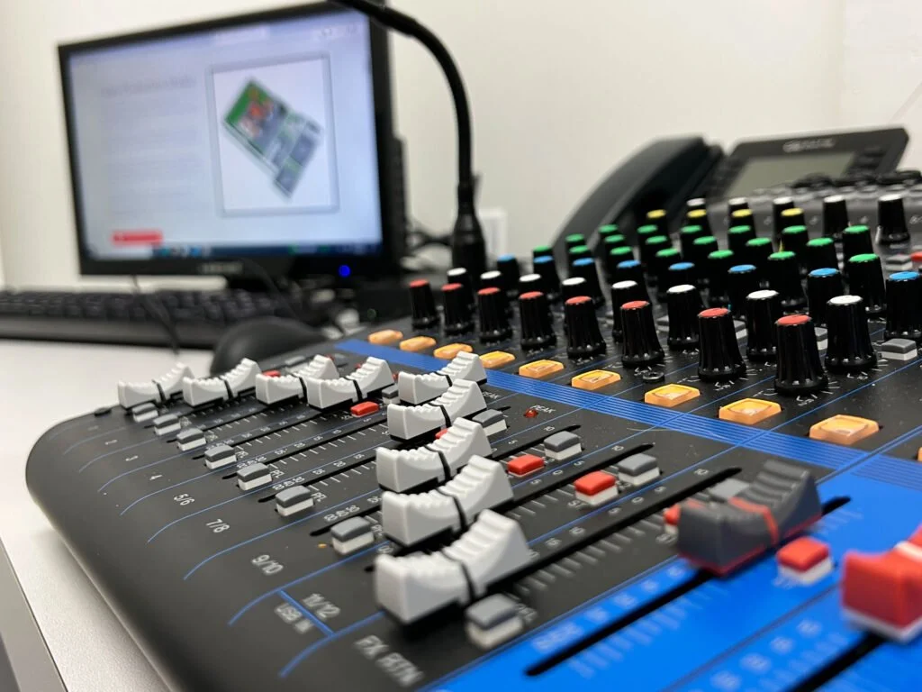 Audio mixing console with sliders and knobs, computer monitor in the background showing a blurred image, office phone on the desk.