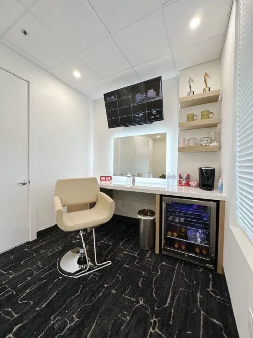 A modern hotel or office break room with a beige swivel chair, built-in mini fridge, shelving with decorative items, coffee maker, and a flat-screen monitor on the ceiling.