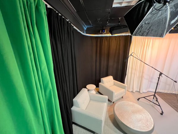 A behind-the-scenes photography or video studio setup featuring a green screen, black and white curtains, two white armchairs, a small round side table, a large umbrella light, and a round woven rug.
