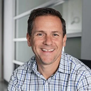 A smiling man in a checked shirt standing outside a house with a glass door.