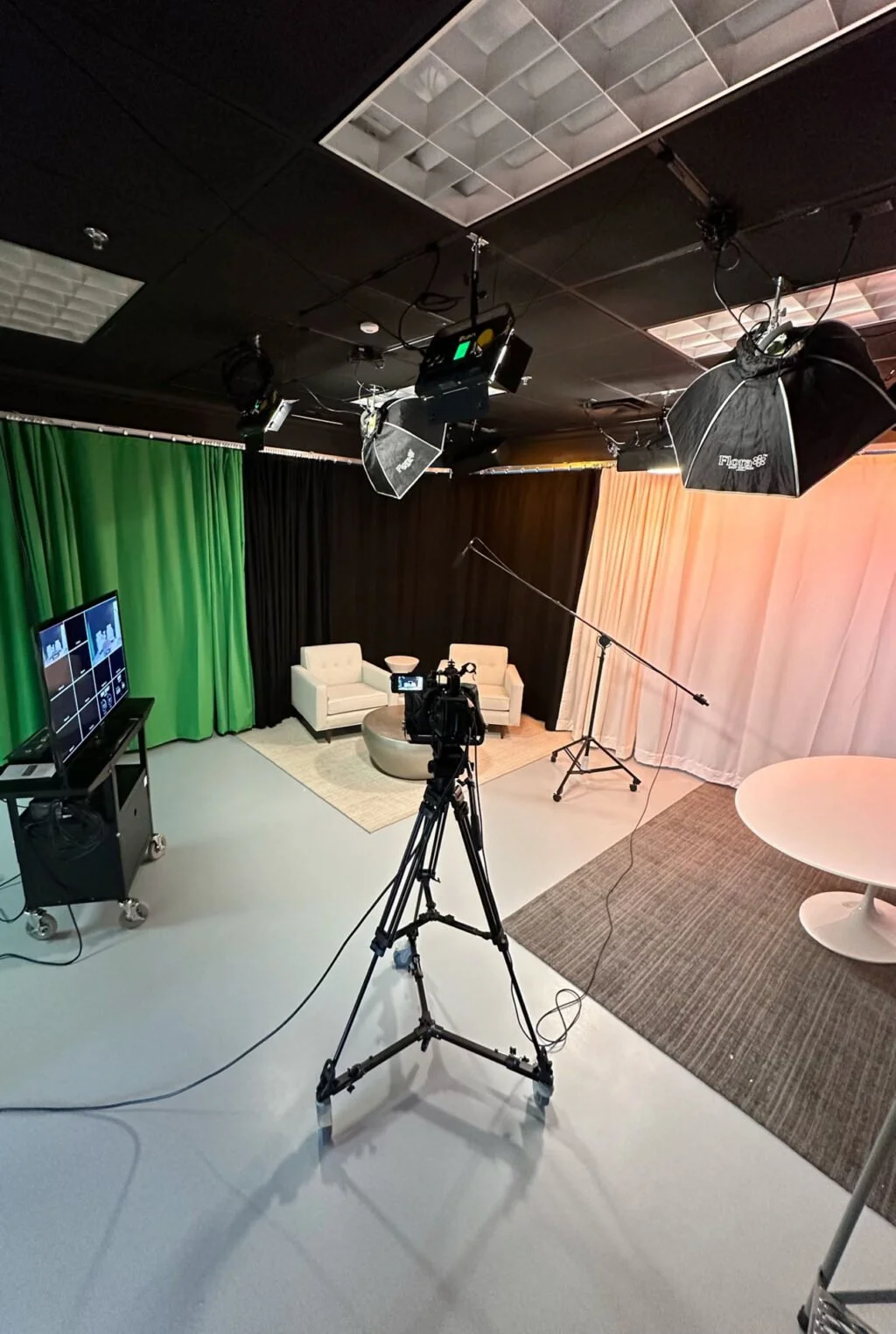 Interior view of a professional video or photo production studio with lighting equipment, camera on tripod, a green screen, black and beige curtains, two white sofas, a TV monitor, and a round table.