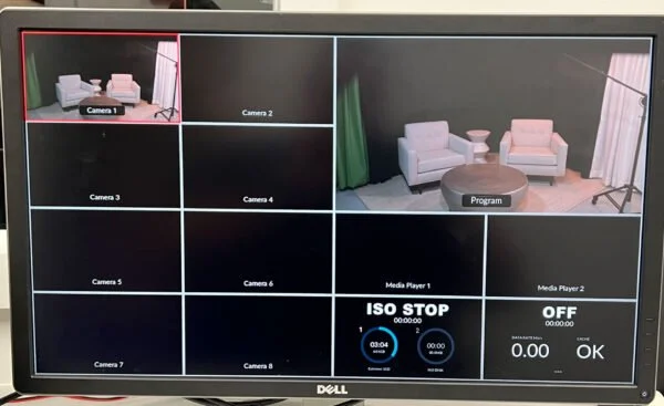 Monitor screen displaying a video conference with multiple camera feeds; main view shows a small seating area with two pink armchairs, a round coffee table, and a floor lamp with a white shade.