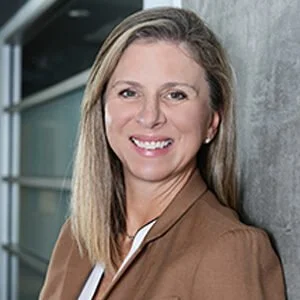 A smiling woman with blonde hair wearing a tan blazer in front of a gray wall