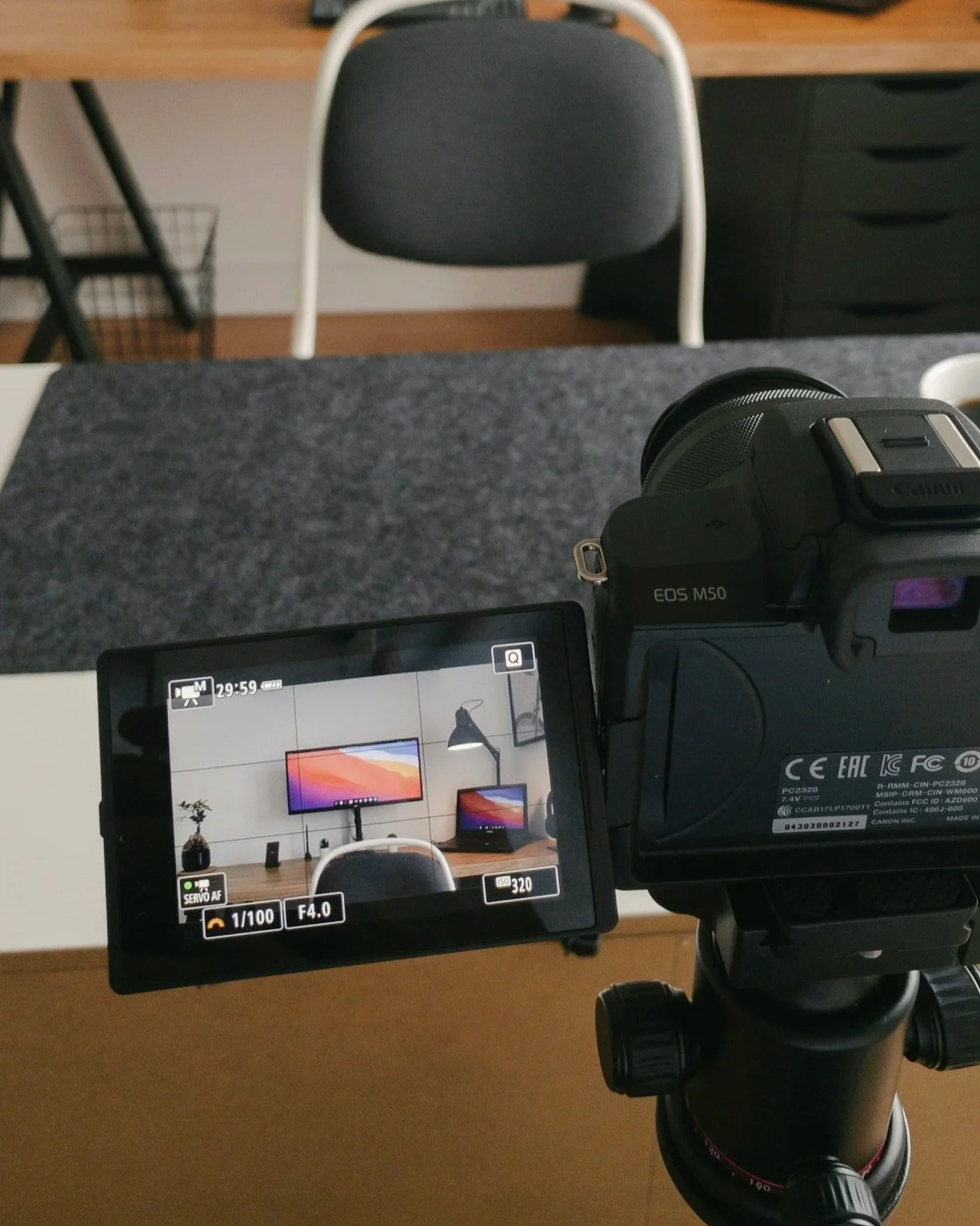 Camera on a tripod capturing a photograph or video of a workspace featuring a desk, chair, three monitors, and a small figurine.