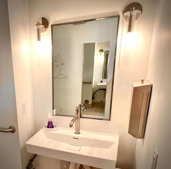 Bathroom with a rectangular mirror above a white sink, wall-mounted lights on each side of the mirror, a soap dispenser on the sink, and a wooden cabinet on the right wall.