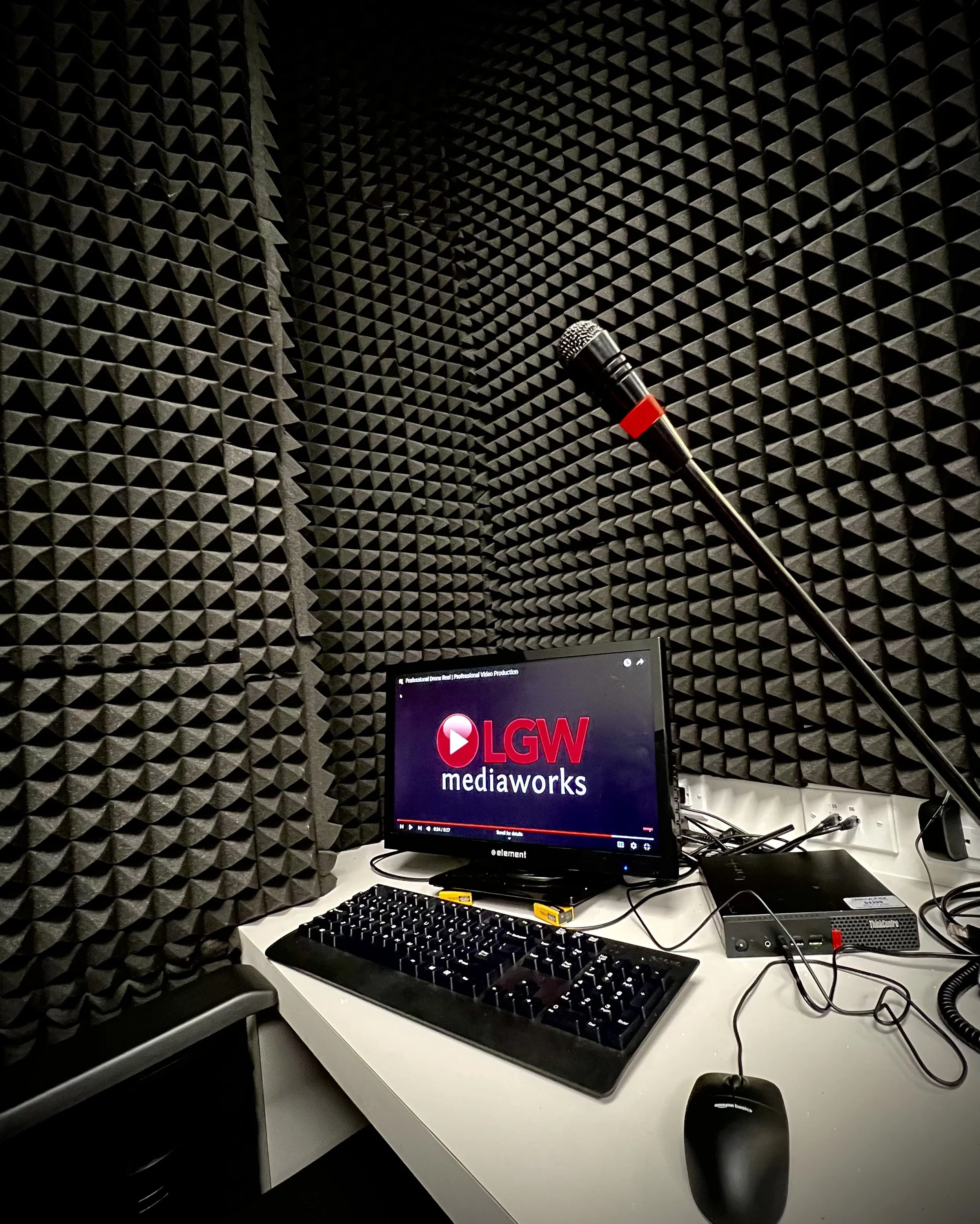 Recording studio with soundproof foam panels, a computer monitor displaying the LGW MediaWorks logo, a keyboard, mouse, and audio equipment on a white desk.