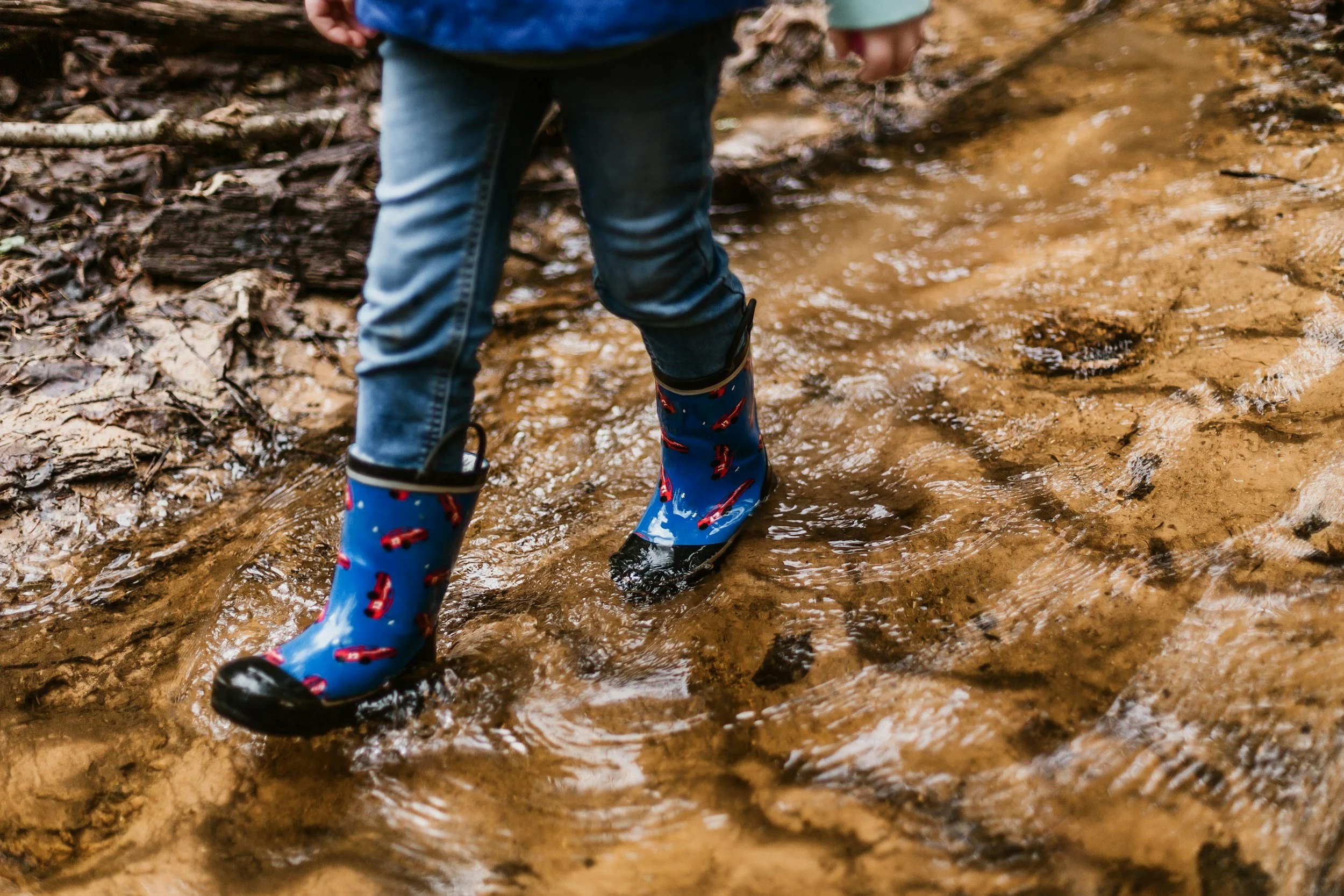 Child wearing blue boots with red lightning bolts, blue jacket, and jeans, standing in muddy water outdoors.