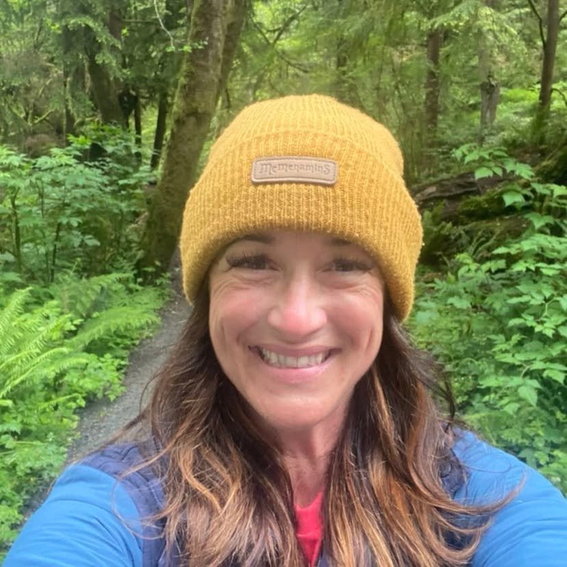 A woman smiling outdoors on a wooded trail, wearing a yellow knit beanie and a blue jacket.