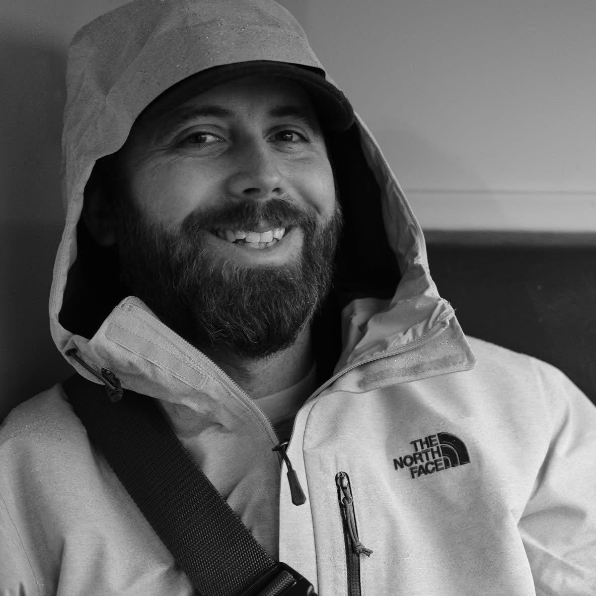A smiling man with a beard, wearing a hooded jacket from The North Face, sitting indoors.