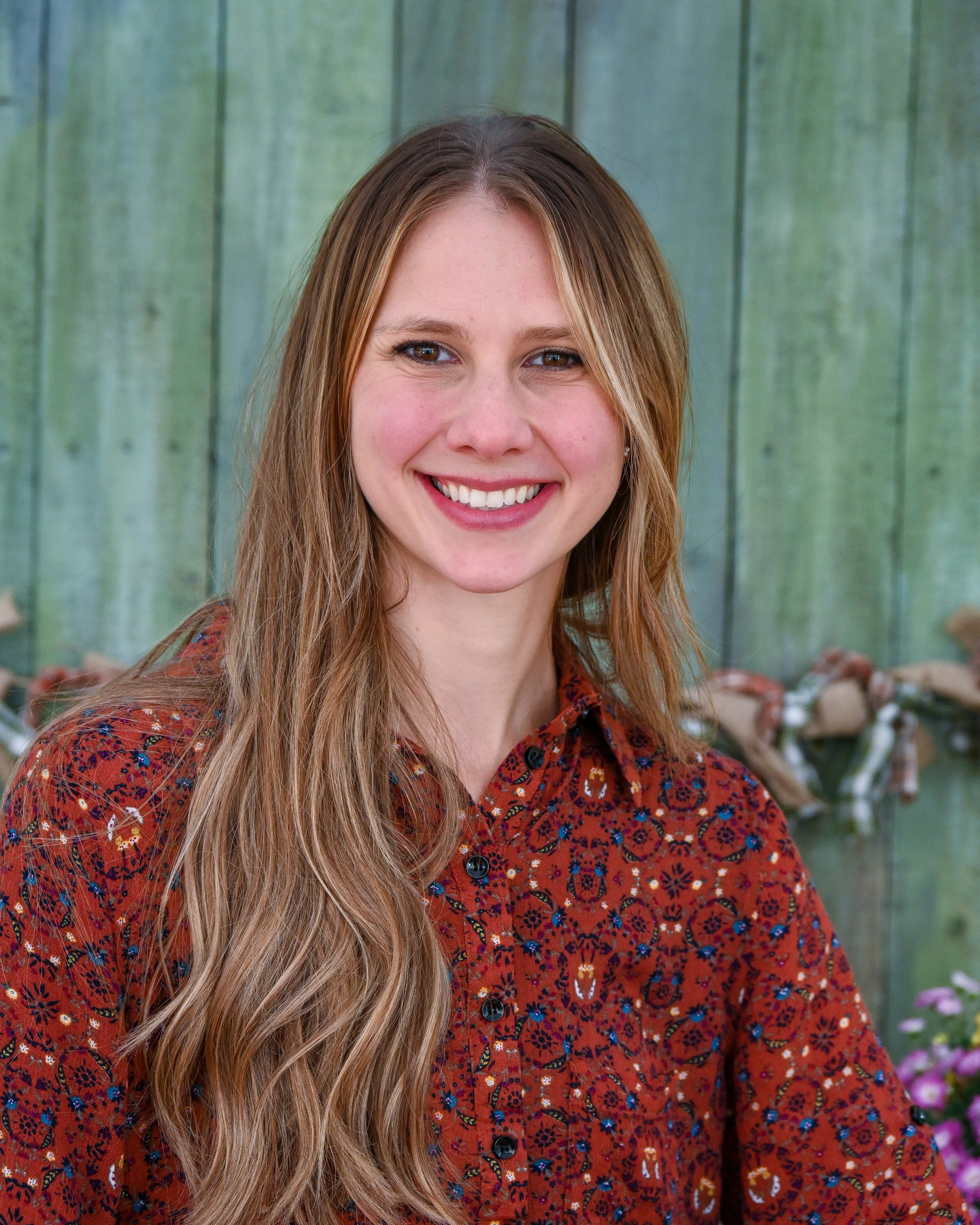 Young woman with long, wavy, light brown hair smiling, wearing a red patterned blouse, standing outdoors in front of a green wooden fence, with flowers in the background.