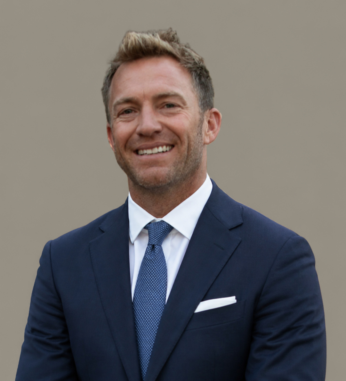 A man in a navy blue suit, white shirt, and blue tie smiling outdoors with a plain beige background.