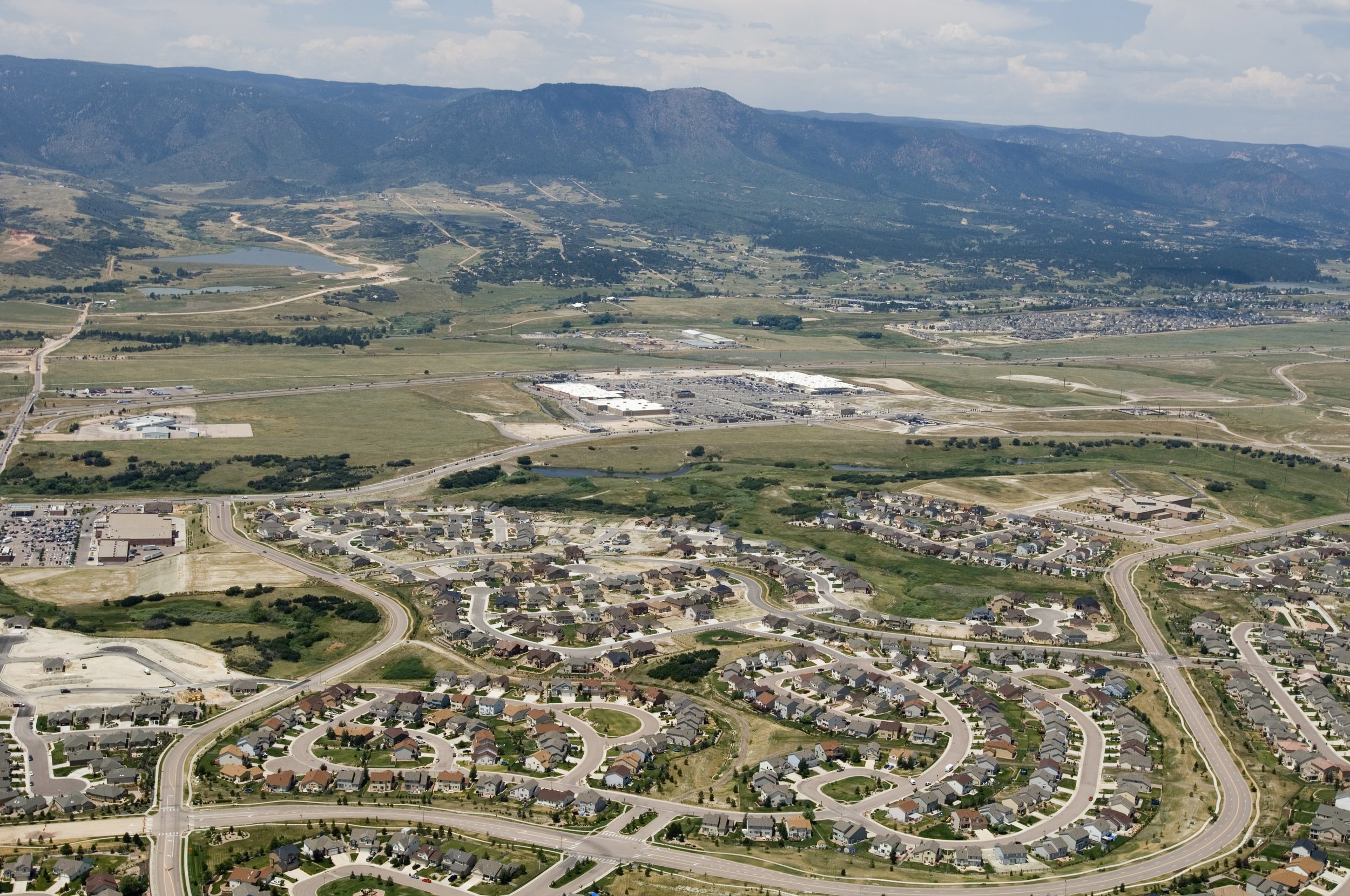 Aerial view of a suburban neighborhood with winding streets, houses, industrial area, farmland, a small lake, and mountains in the background under a partly cloudy sky.