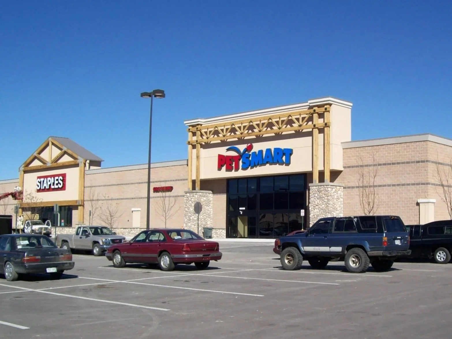 View of a shopping plaza with PetSmart and Staples stores, a parking lot with several parked cars, and a clear blue sky.