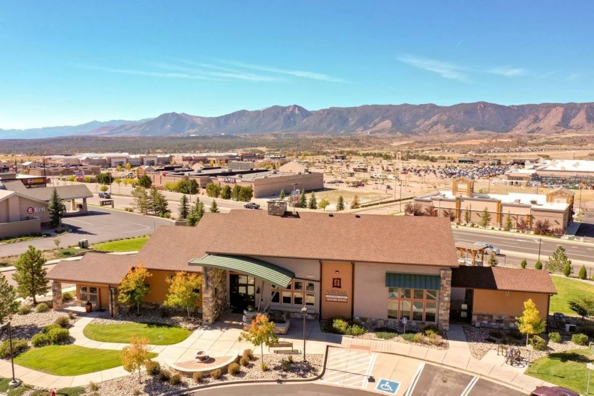 A small building with a green awning, a landscaped front yard, and a parking lot with handicap spots, with a shopping mall and distant mountains under a clear blue sky in the background.