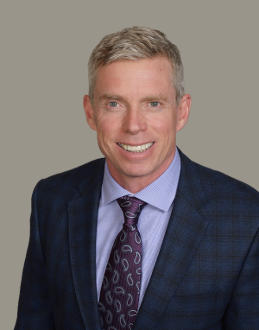 Portrait of a smiling middle-aged man in a blue suit, light blue shirt, and patterned tie against a plain gray background.