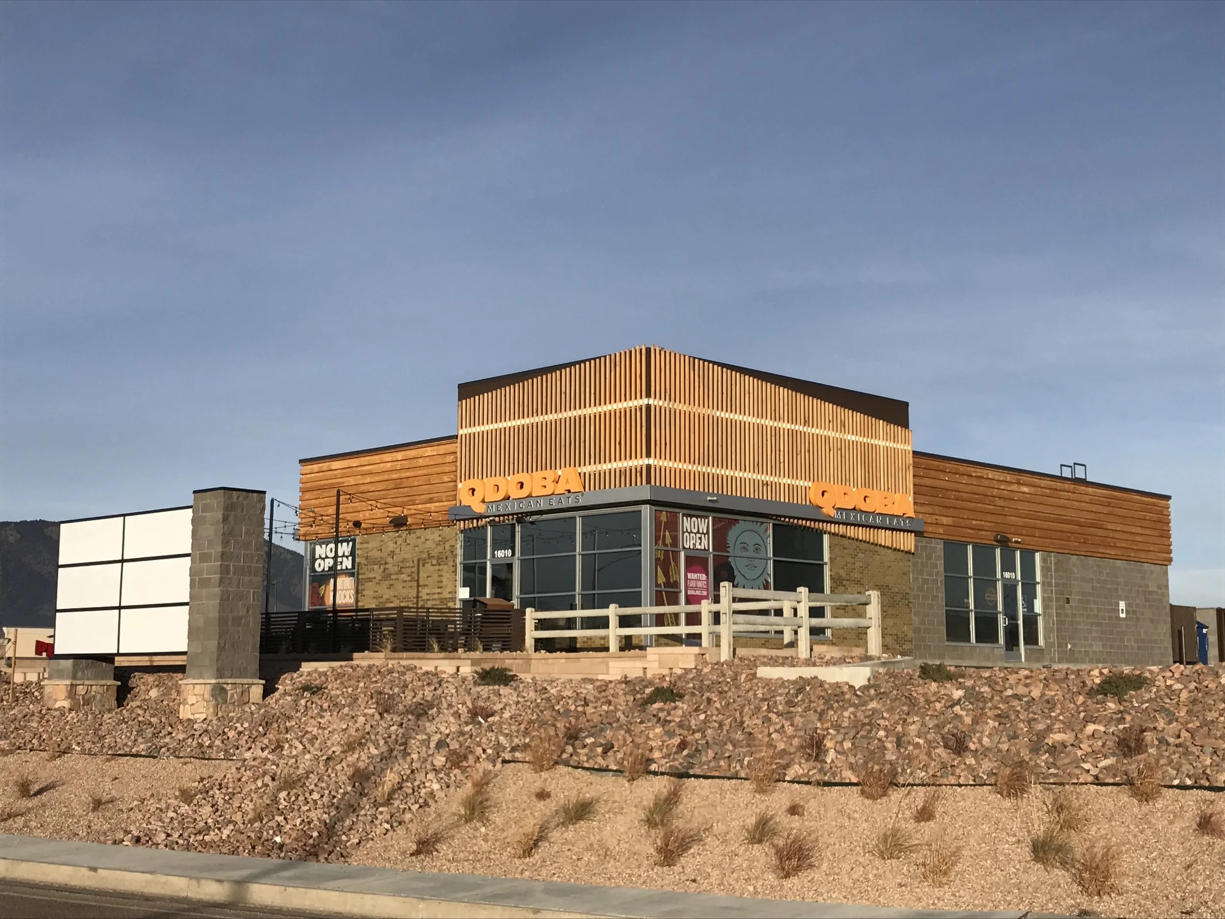 Modern building with wood paneling and large glass windows, signage indicating it is a Mexican restaurant called "QDOBA," located at 16010. The building has a distinctive roofline with geometric shapes, and the surrounding area has rocks and sparse desert plants. The sky is clear with a few clouds.