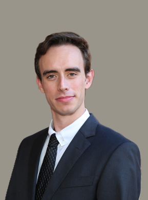 Portrait of a young man in a dark suit and tie, smiling slightly, against a plain background.