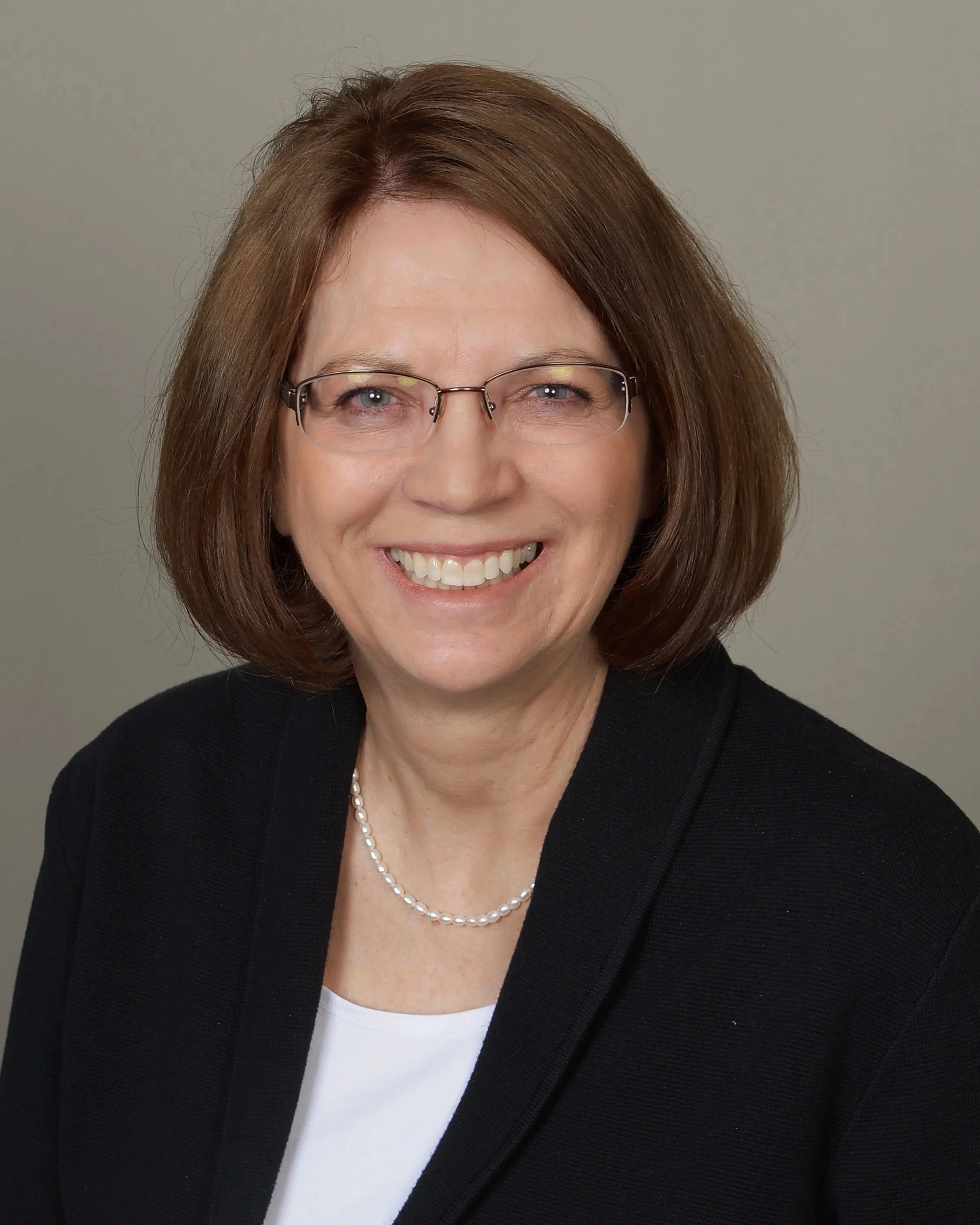 Close-up of a smiling woman with shoulder-length brown hair, glasses, wearing a pearl necklace, black blazer, and white top, neutral background.