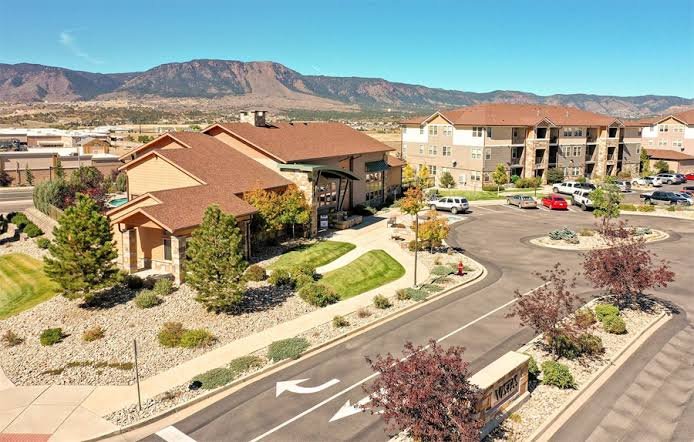 View of a residential apartment complex with landscaped greenery, parking lot, and distant mountains.