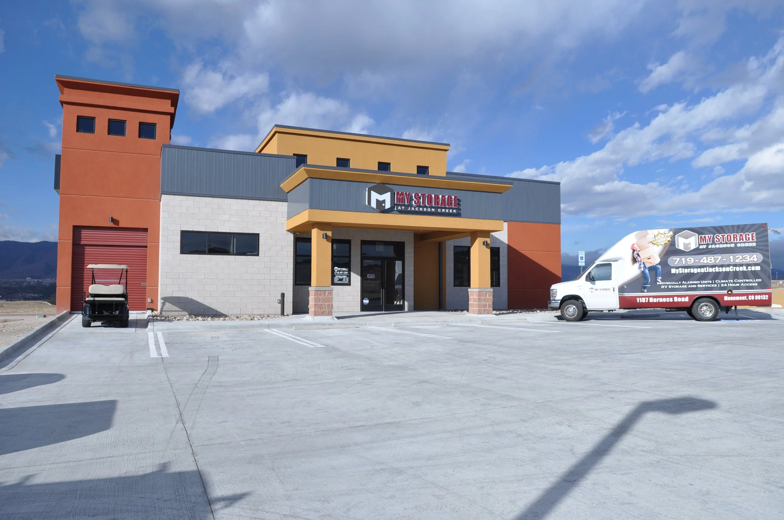 A building with a sign that reads "My Storage at Jackson Creek" and a parking lot with a golf cart and a delivery truck. The building has multiple sections with colorful exterior walls and a small covered entrance.