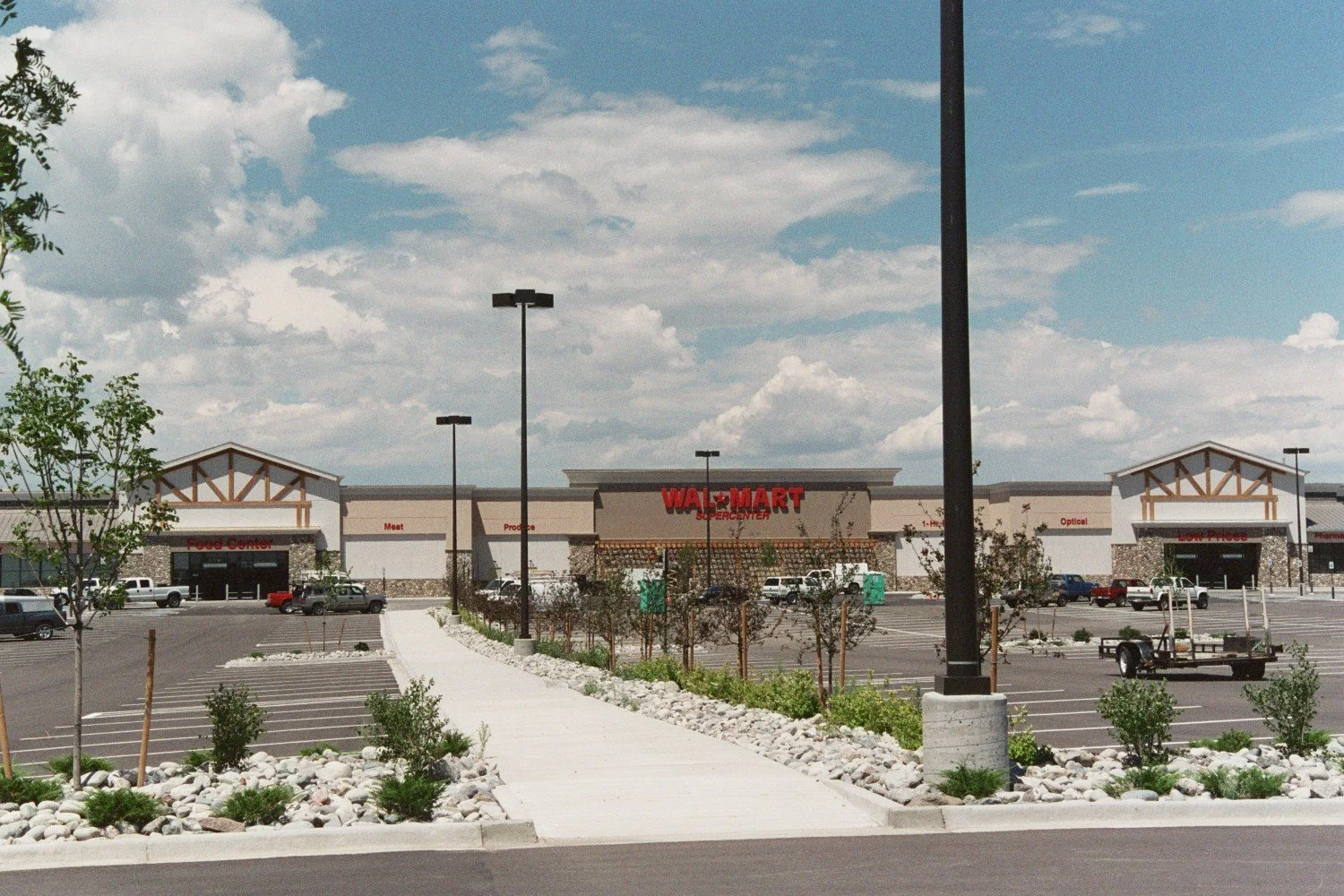 Exterior of a new Walmart Supercenter with a large parking lot, some trees, and a sidewalk. The Walmart sign is prominent, and there are a few parked cars and empty parking spaces.