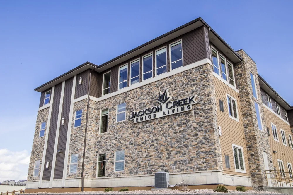 Exterior of Jackson Creek Senior Living apartment building with stone and siding facade, multiple windows, and a sign reading 'Jackson Creek Senior Living' on the front.