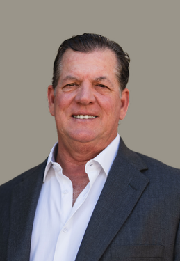 A middle-aged man with dark hair, wearing a dark suit jacket and white shirt, smiling against a gray background.