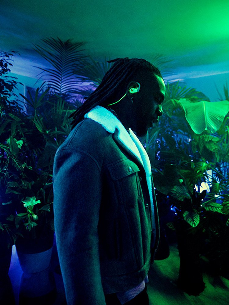 A man with dreadlocks wearing a denim jacket with a white fleece collar, standing among green and purple plants illuminated by green and blue lighting.