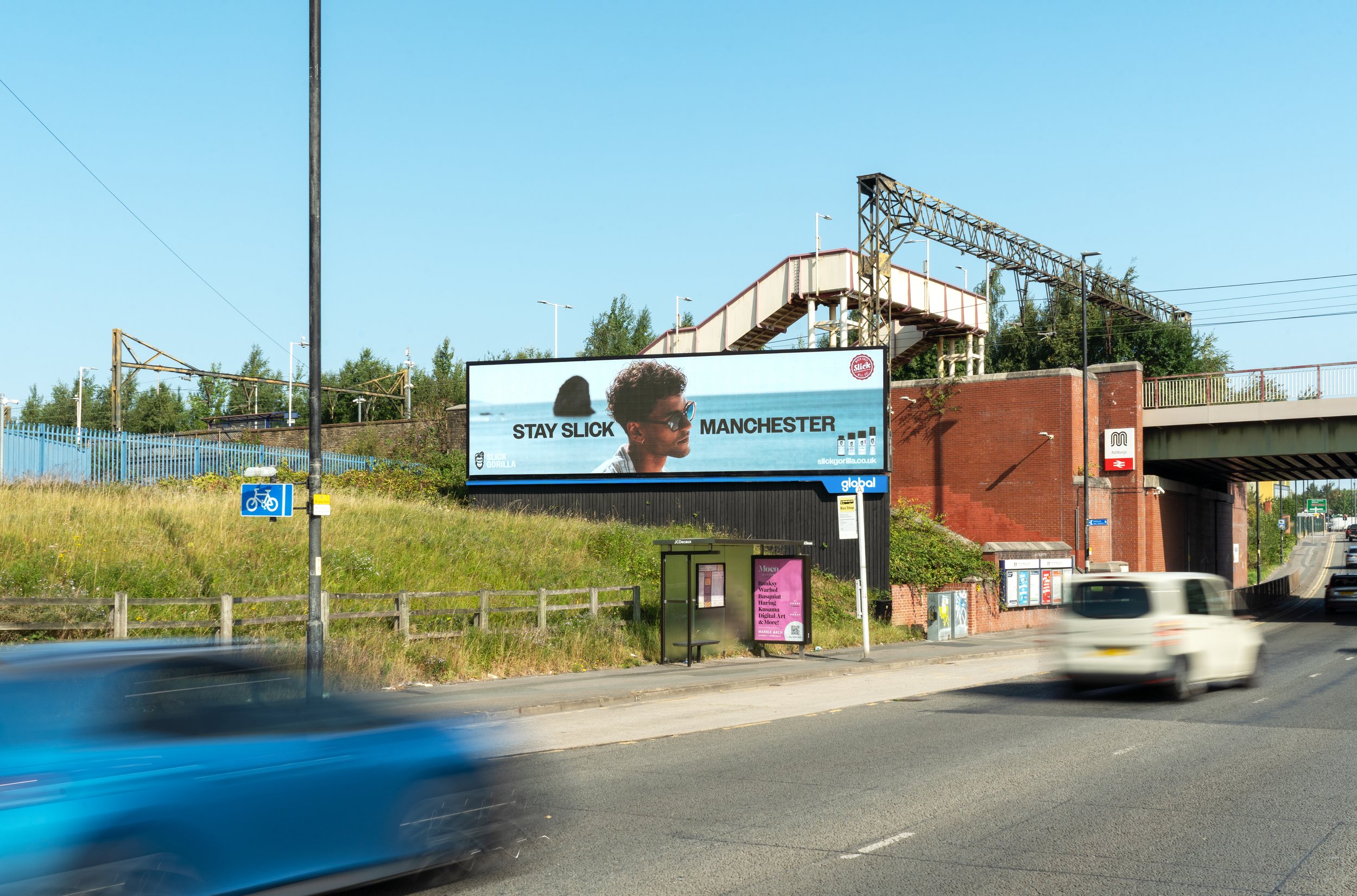 A highway overpass with a large billboard advertising Slick Gorilla, featuring a man wearing sunglasses and the text 'Stay Slick Manchester'. In the foreground, a moving blue car and a blue bicycle lane sign are visible, with some greenery and trees in the background.
