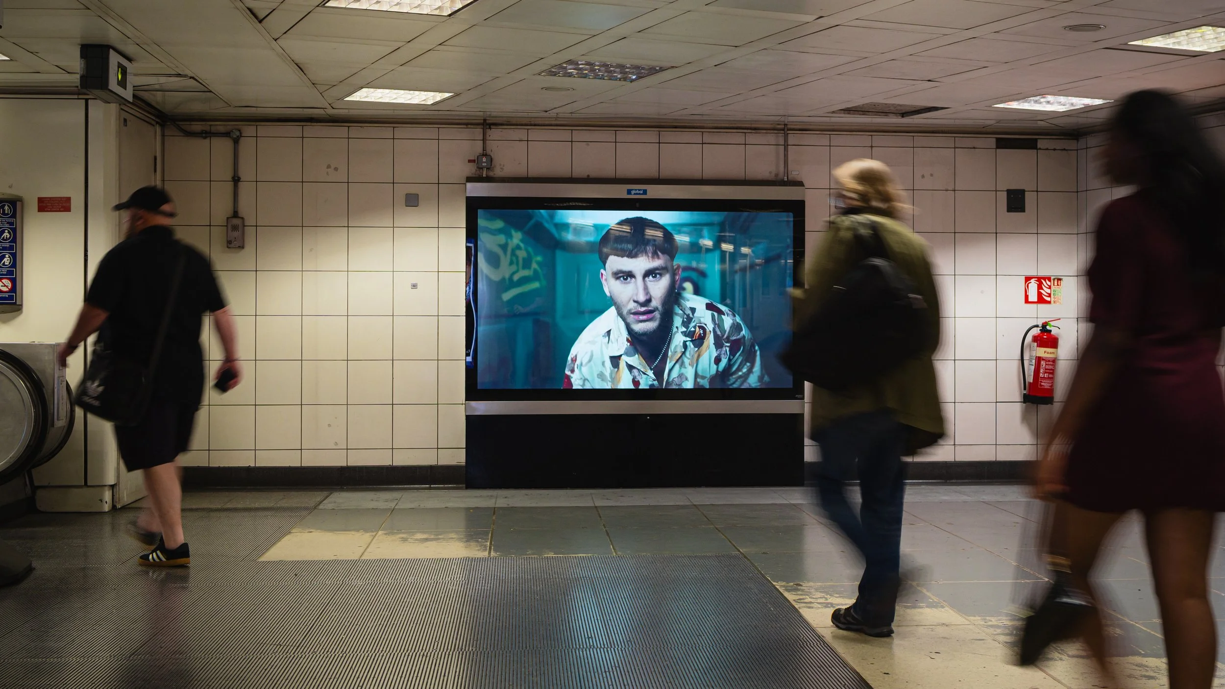 A subway station with a large advertisement screen showing a man's face. Several people are walking past the screen, some blurred with motion, and the station has tiled walls and a fire extinguisher on the right side.