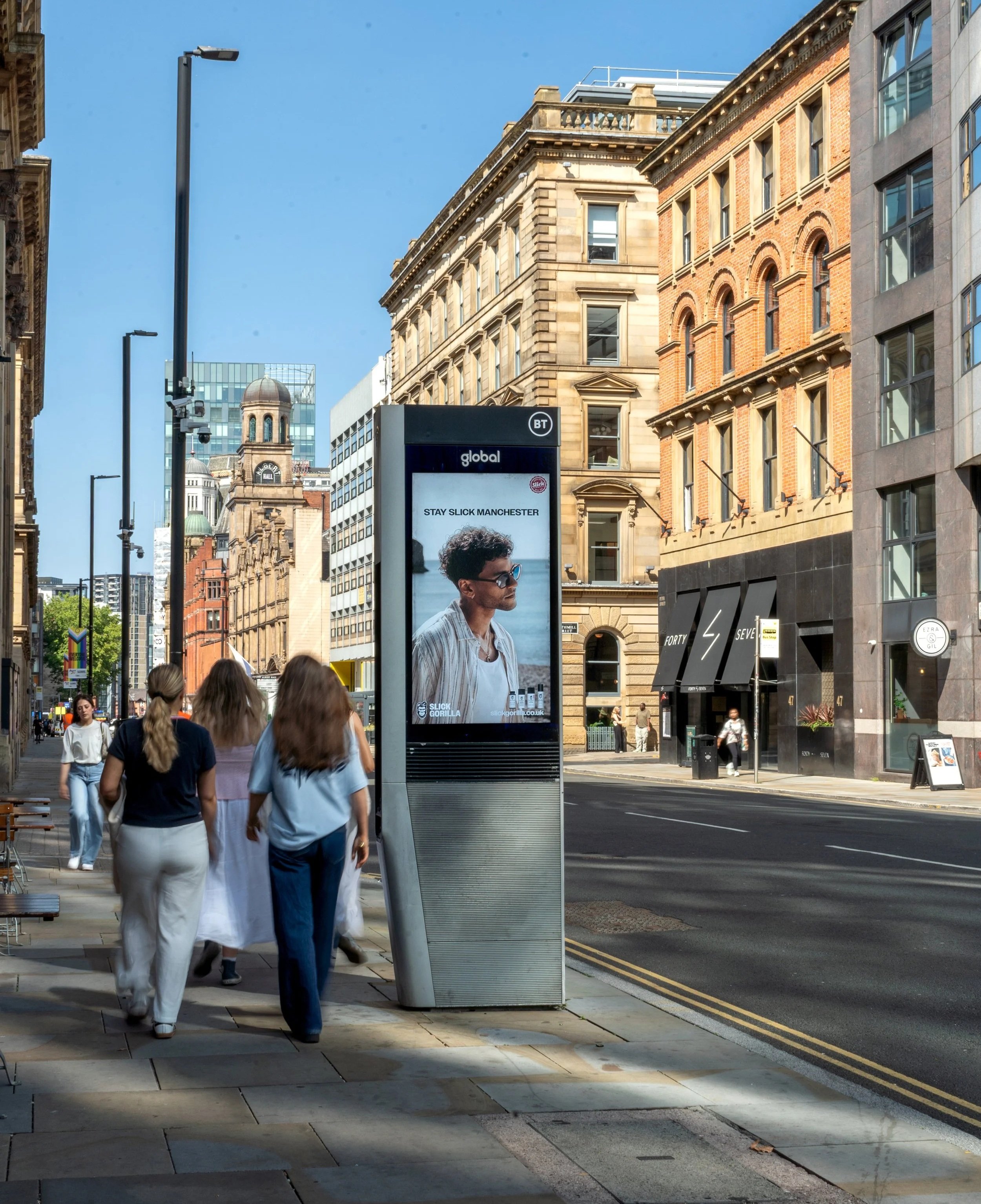 People walking on a city sidewalk near a street with buildings and streetlights, a digital billboard displaying an advertisement, and a clear blue sky.