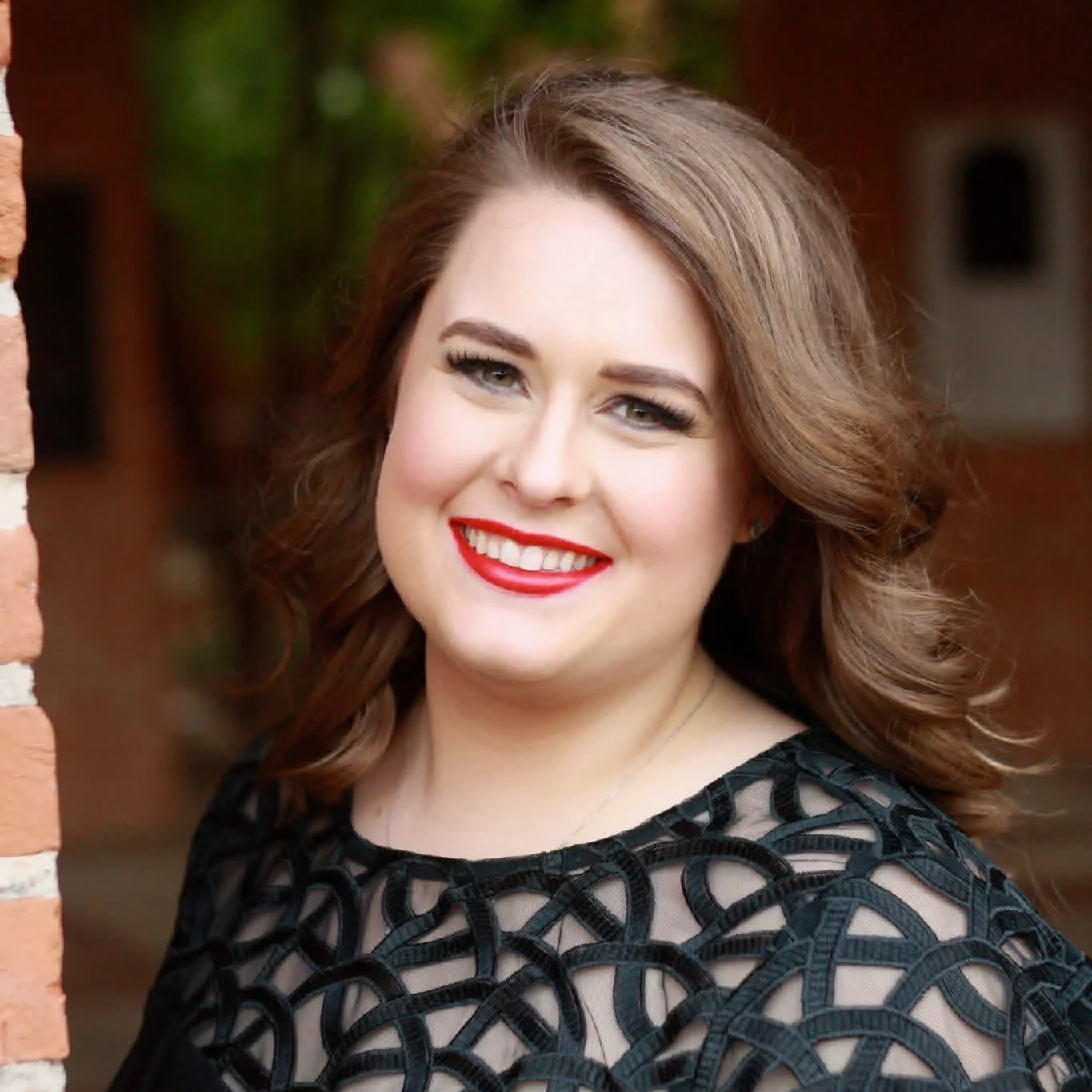 Portrait of a woman outdoors with a blurred background, smiling, with wavy brown hair, red lipstick, makeup, wearing a black dress with circular cutouts.