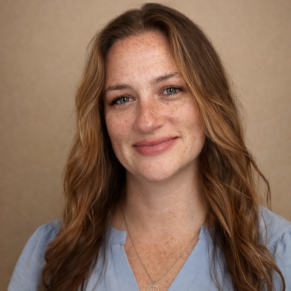 A smiling woman with long, wavy red hair, green eyes, and freckles, wearing a light blue top and a delicate necklace, against a neutral beige background.