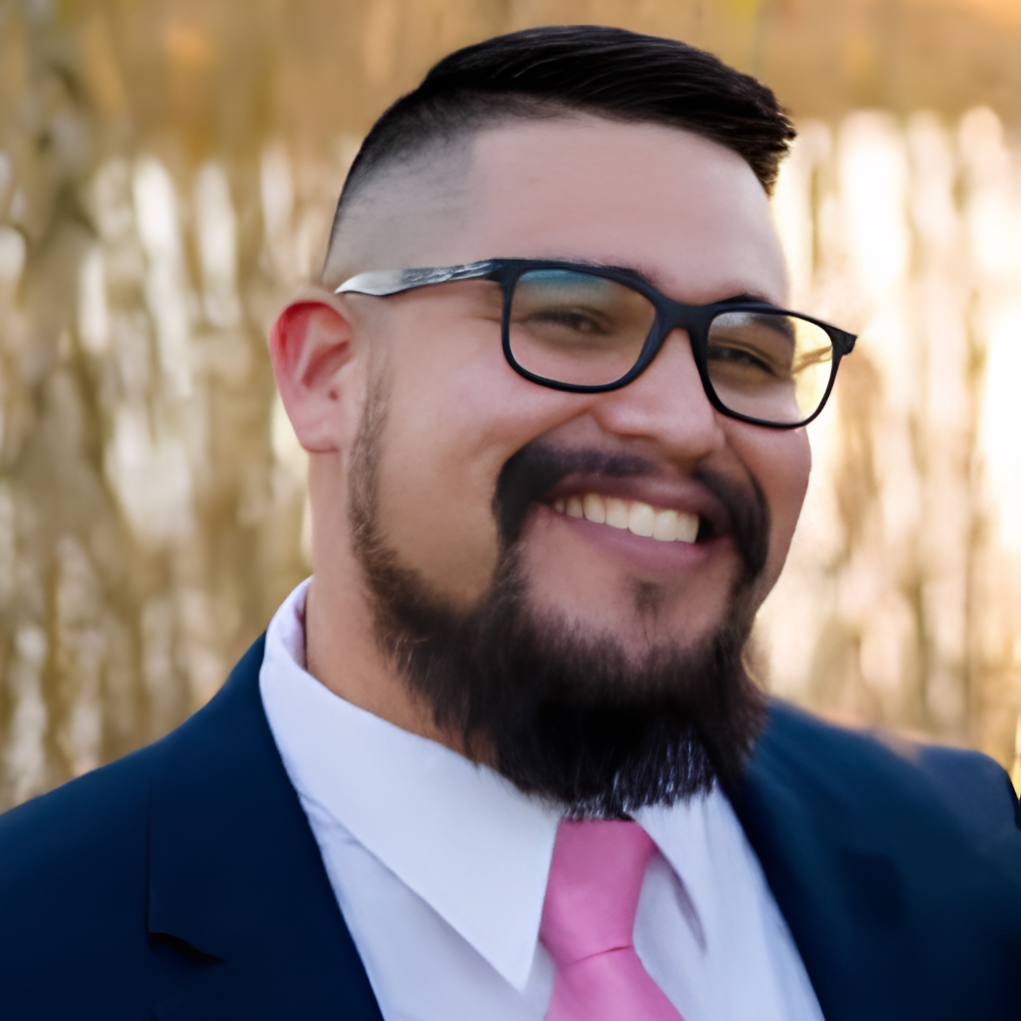 A smiling man wearing glasses, a navy blue suit, a white shirt, and a pink tie outdoors during golden hour.