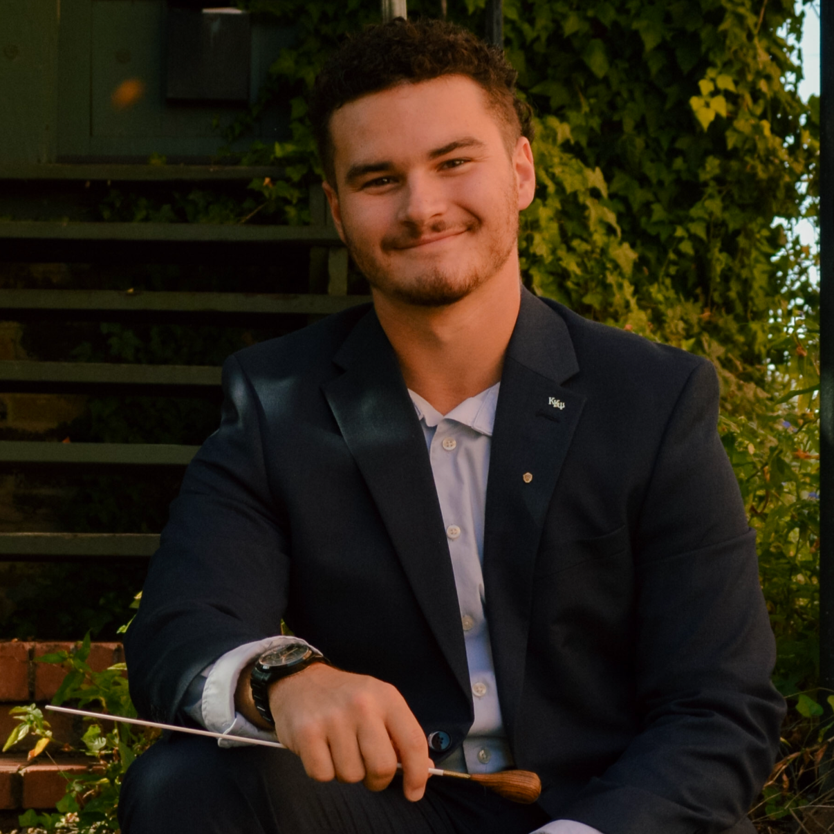 A young man with short curly hair, facial hair, and a smile, dressed in a dark blazer and light shirt, sitting outdoors with greenery and stairs in the background, holding a conductor's baton.