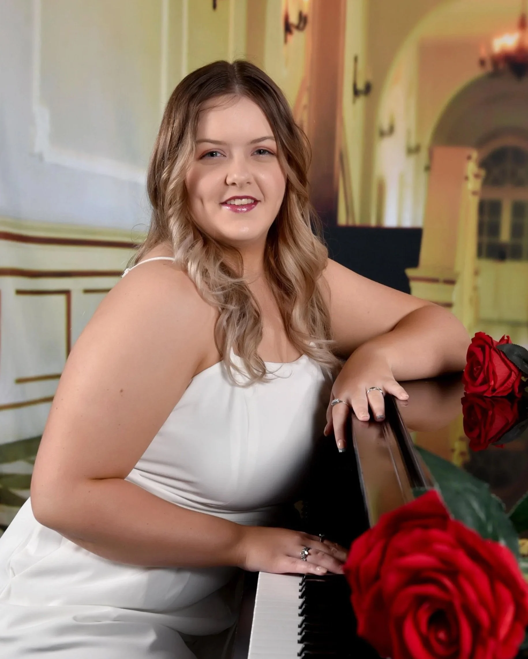 A woman with light brown hair sitting at a black and white piano, smiling, with a bunch of red roses in front of her.
