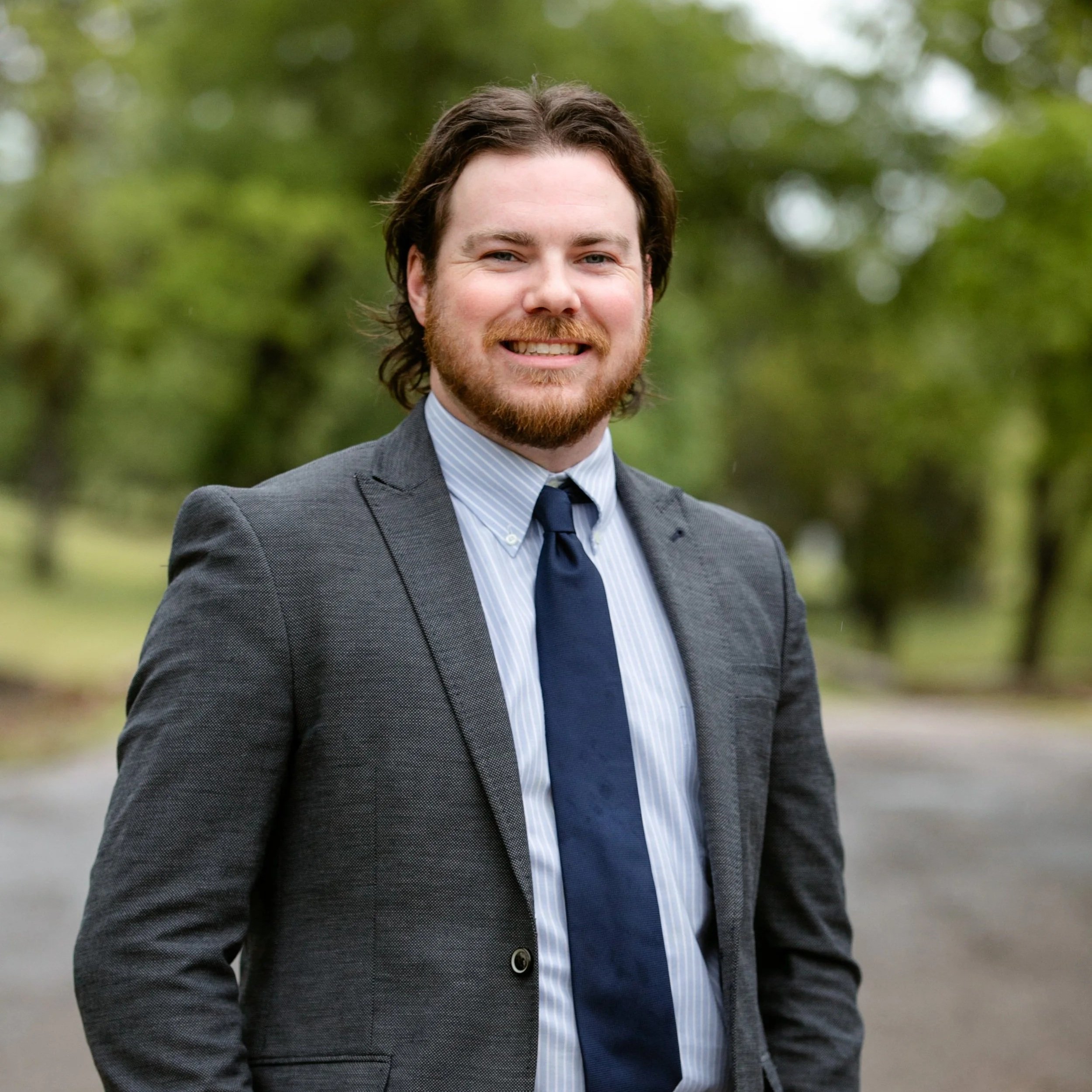 A man with brown hair, a beard, dressed in a gray checkered suit, white and blue striped shirt, and a dark blue tie, standing outdoors with a blurred green background, smiling at the camera.