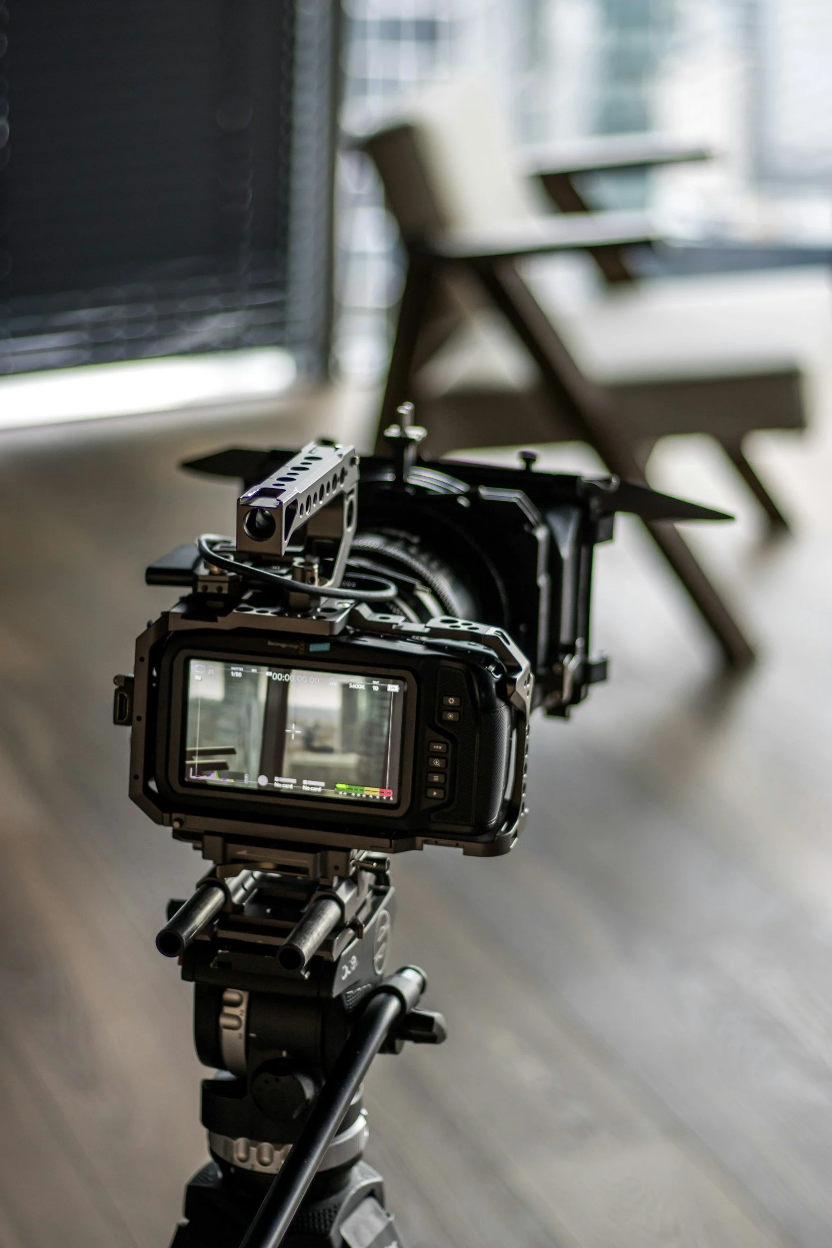 A professional camera mounted on a tripod with a matte box, set up for filming or photography, in a room with a modern design and large windows in the background.