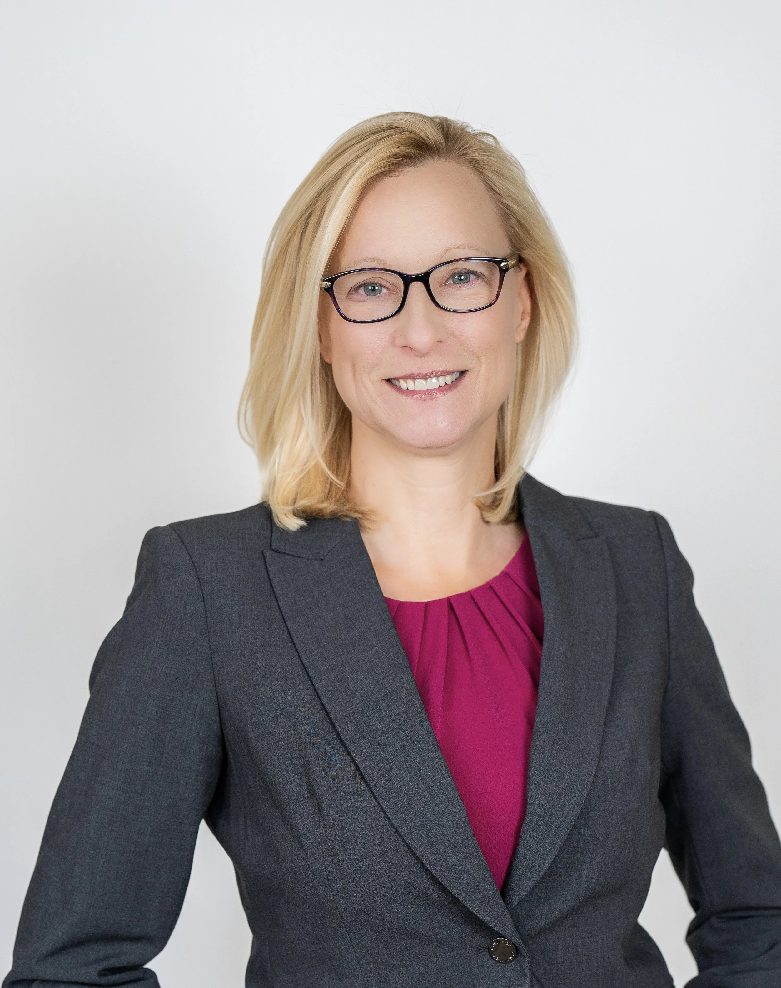 A woman with blonde hair, wearing glasses, a dark gray blazer, and a magenta top, smiling against a plain white background.