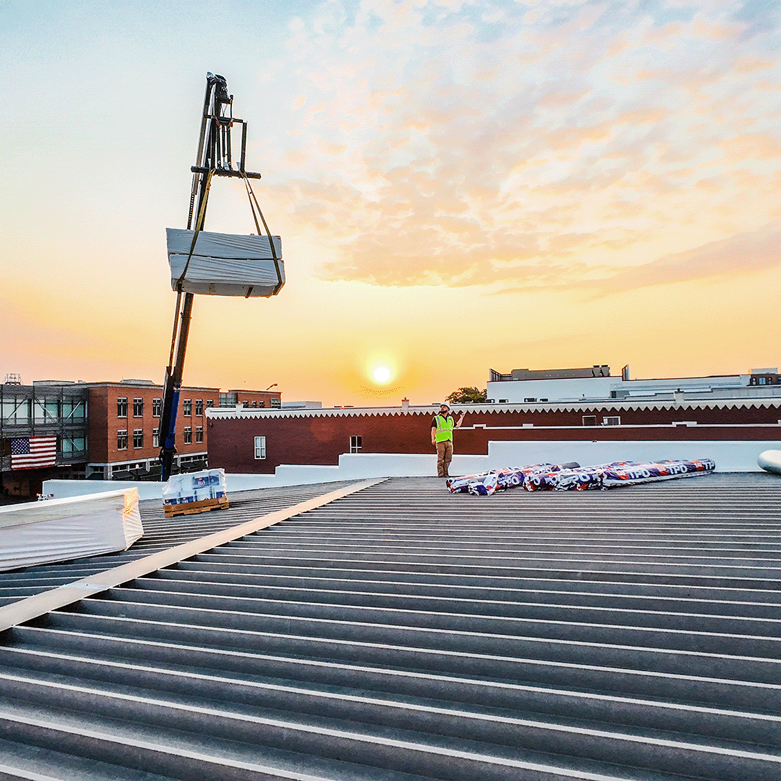 Worker on a rooftop during sunset, with roofing materials and a crane lifting supplies.