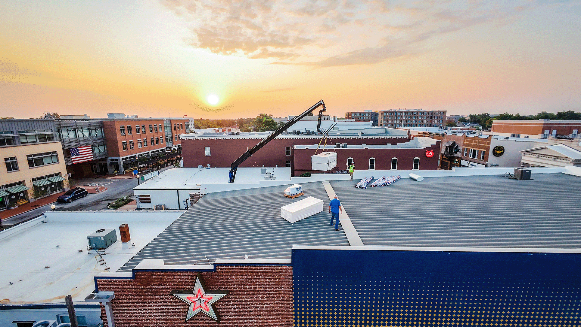 View of a city rooftop at sunset, with workers installing equipment, a crane lifting a box, and various buildings in the background.